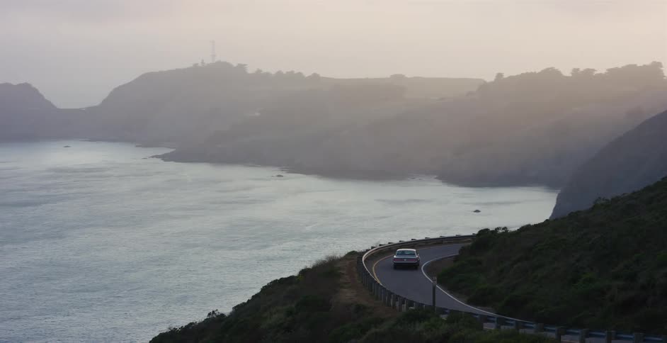 A vintage car travels down a winding, foggy coastal highway overlooking the ocean at sunset