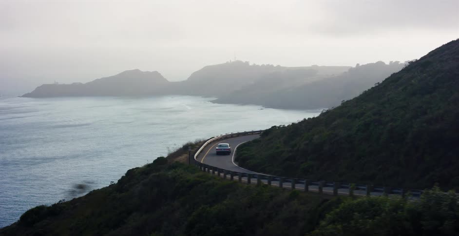 An aerial shot follows a classic car as it drives along a scenic, winding coastal road on a very foggy day