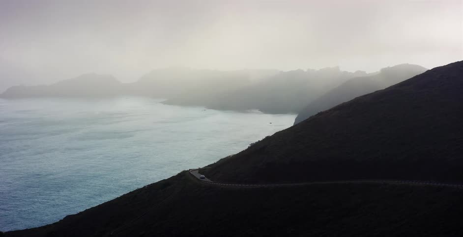 An aerial view of a car driving along a winding coastal highway as it becomes enveloped in thick fog