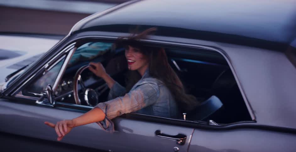 A carefree woman with long hair blowing in the wind smiles while driving a vintage muscle car in slow motion