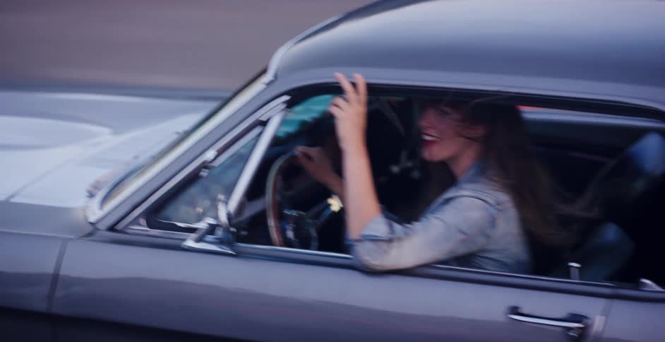 A happy woman with her hair blowing in the wind drives a classic muscle car while flashing a peace sign out the window