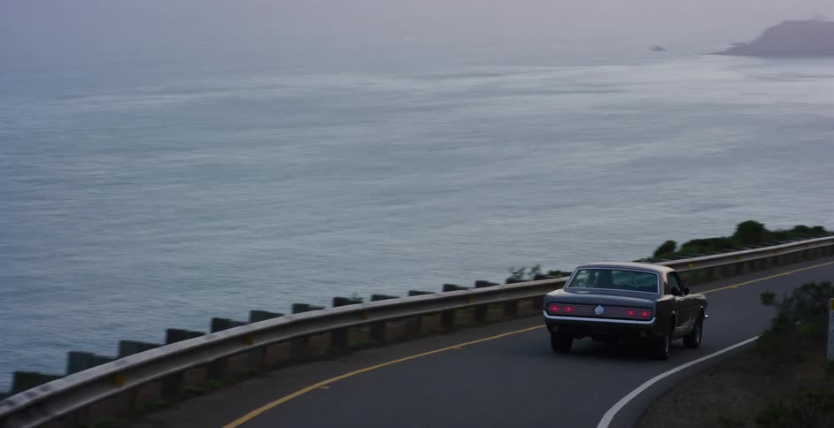 A vintage muscle car drives along a scenic, winding coastal road overlooking the ocean on a foggy evening
