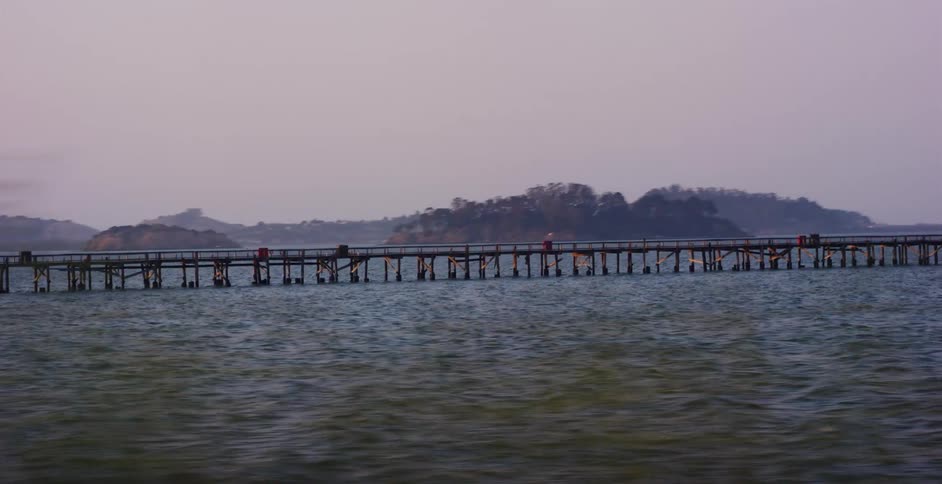 A scenic side-view tracking shot captures a long wooden pier stretching across the calm ocean at dusk