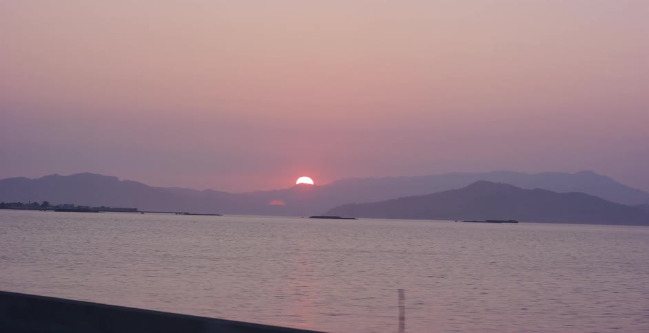 A scenic drive alongside a tranquil lake reveals a beautiful pink and purple sunset dipping behind distant mountains
