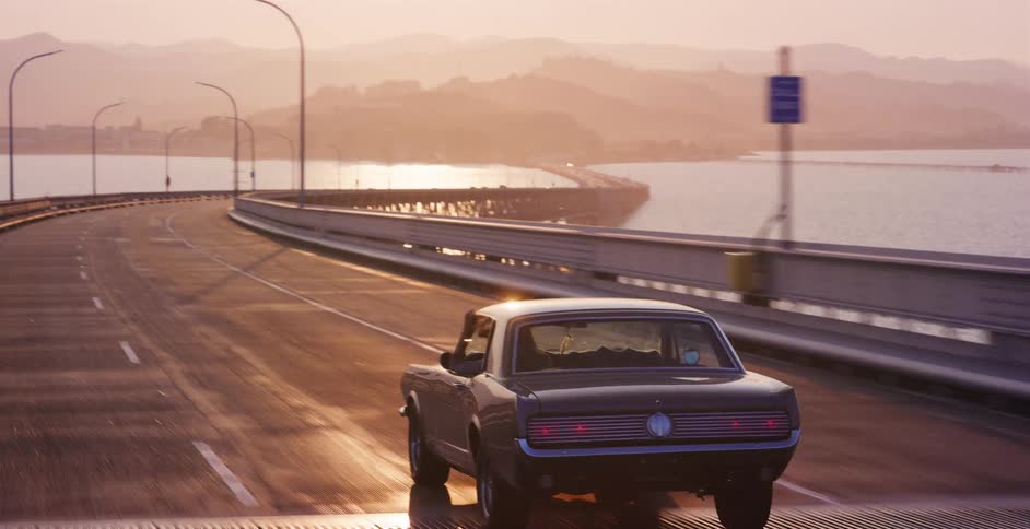 A vintage black muscle car drives along a long, winding coastal bridge bathed in the warm, golden light of a beautiful sunset