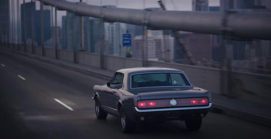 A sleek vintage muscle car drives across a bridge with a modern city skyline in the background at dusk