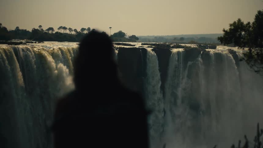 A slow-motion, over-the-shoulder shot captures the silhouette of a person admiring the awe-inspiring Victoria Falls at sunset