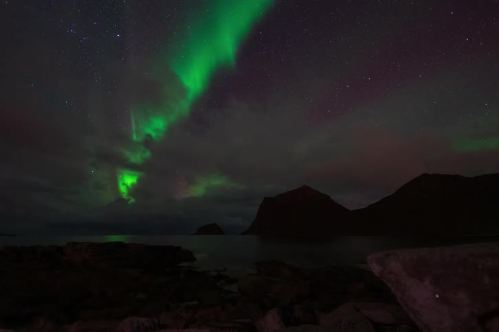 Vibrant green Northern Lights dance across a cloudy, star-filled sky in a stunning time-lapse over a rocky coastal landscape