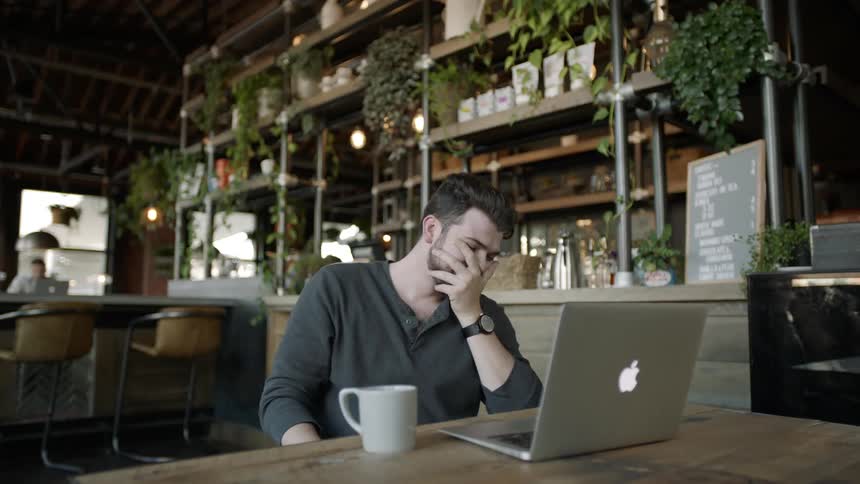 Man sitting at table in coffee shop looking at laptop screen and laughing