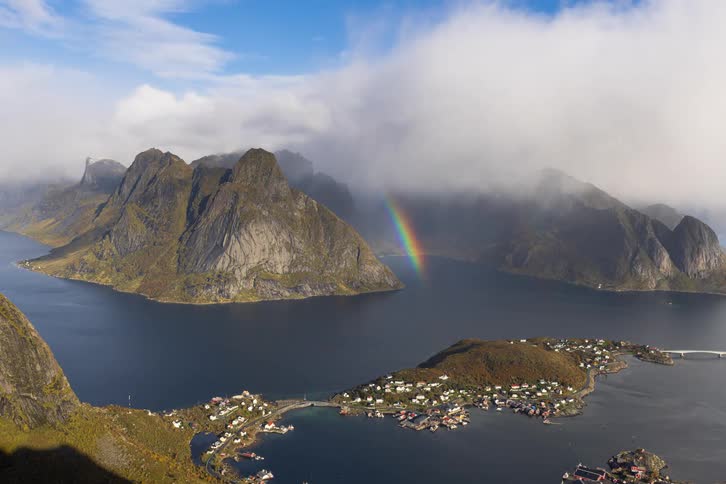 A stunning view from above reveals a bright rainbow emerging from the clouds over the iconic mountains and fishing village of Reine, Lofoten Islands