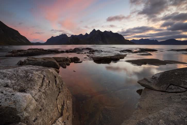Slow dolly time-lapse reveals a majestic sunset over a rocky fjord, with mountains and clouds reflecting in the tranquil water