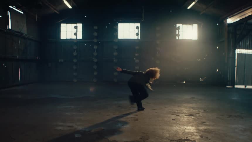 A single dancer with an afro performs an energetic and expressive solo dance in a large, empty, backlit warehouse in slow motion
