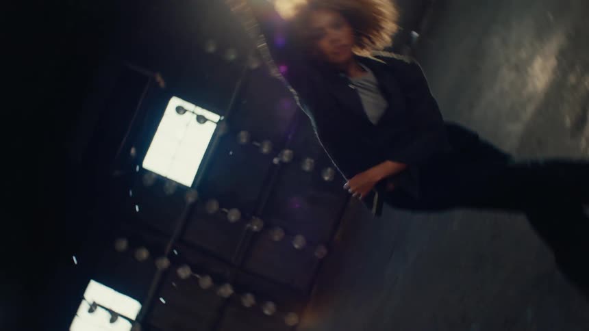 A stylish dancer with a blonde afro floats backwards in slow motion before striking a pose in a vast, sunlit warehouse