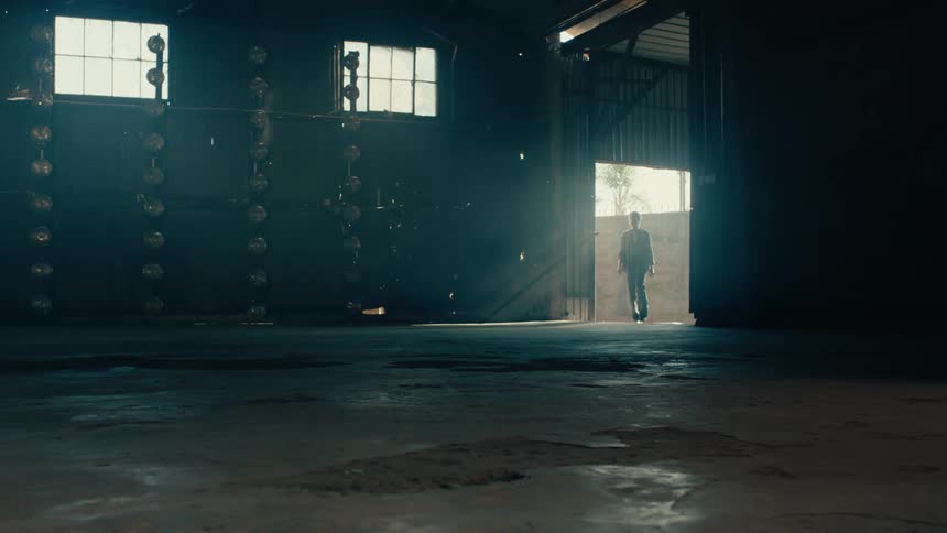 A man opens a door and walks into a large, dark, and empty warehouse with light beams shining through the dust