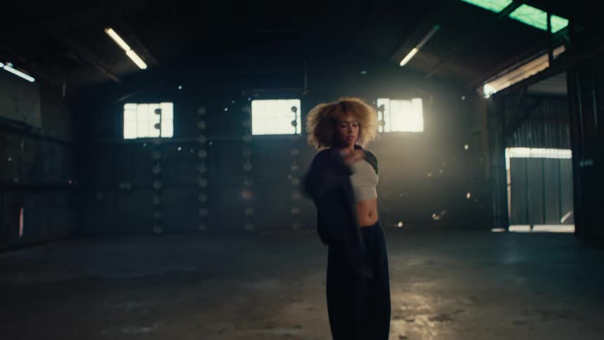 Slow motion shot of a stylish woman with an afro dancing expressively in a large, empty, backlit warehouse