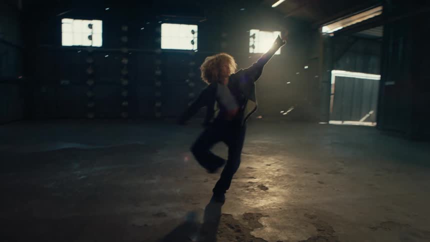 A stylish dancer with voluminous curly hair performs an expressive solo routine in slow motion inside a vast, dark warehouse with dramatic backlighting