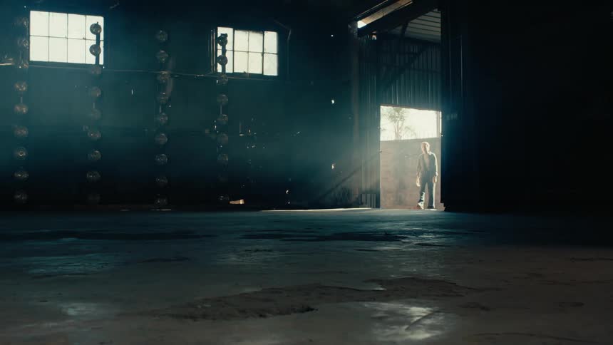 A mysterious man enters a dark, dusty, and smoky warehouse through a brightly lit door, creating a cinematic and dramatic scene