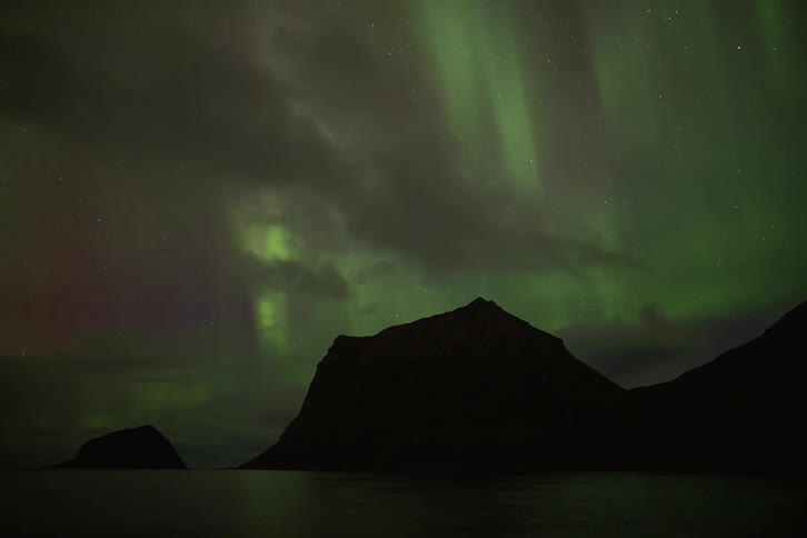 A stunning time-lapse captures the vibrant green Aurora Borealis dancing in the night sky above silhouetted mountains and calm water