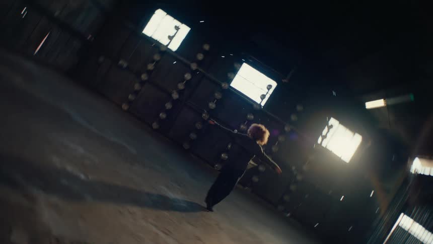 Slow motion vertical shot of an expressive woman with an afro performing a contemporary dance in a vast, sunlit industrial warehouse