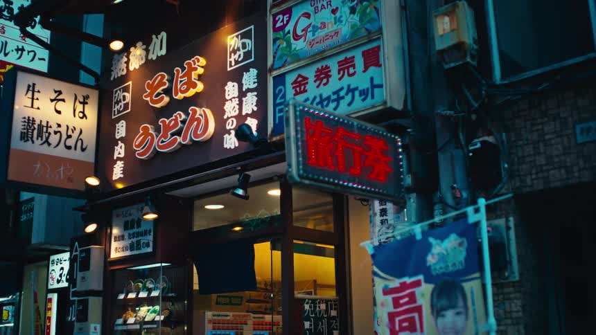 Glowing Japanese signs for a noodle shop illuminate a dark city street as people walk by at night