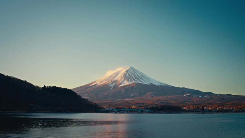 A serene, wide shot shows the majestic, snow-capped Mount Fuji rising above a calm lake during golden hour