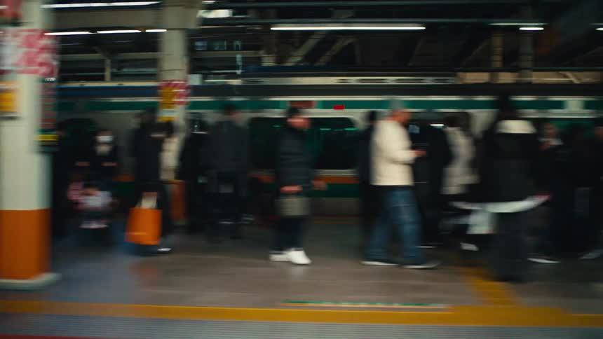 A motion-blurred view from a moving train captures the hurried commuters on a busy platform in a Japanese station