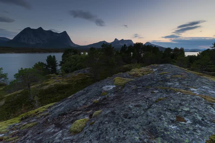 A beautiful timelapse from a rocky, mossy outcrop shows the last light of day fading over a tranquil lake and distant mountains