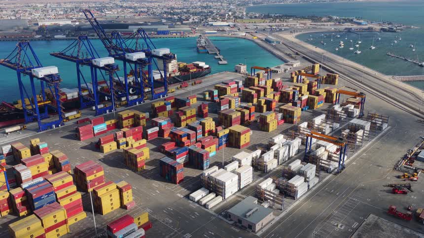 A static aerial view of a busy shipping port with colorful containers, massive cranes, and a cargo ship docked in the harbor