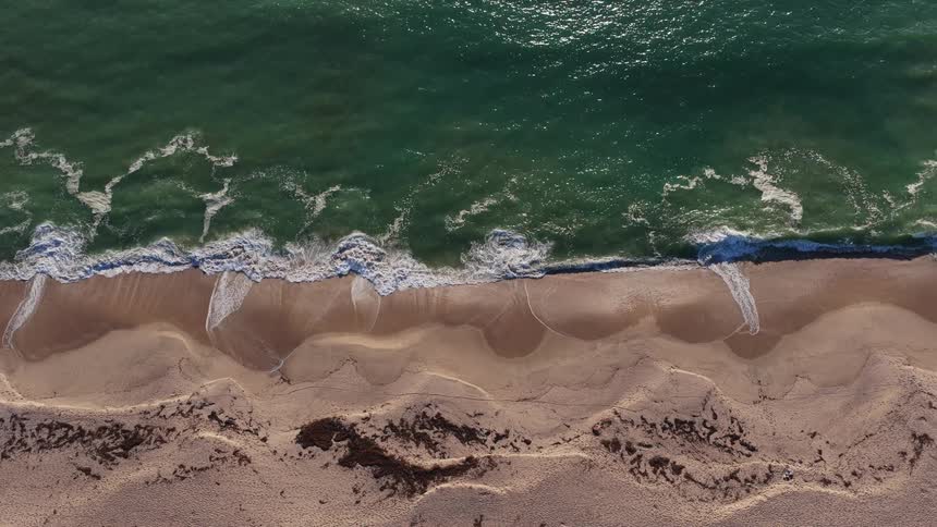 A mesmerizing top-down aerial view shows gentle turquoise waves washing over a serene sandy beach in slow motion