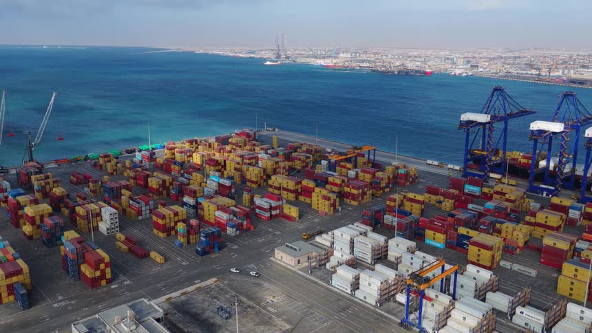 A vast container yard at a bustling seaport, filled with colorful shipping containers next to the ocean, captured in a wide static aerial shot