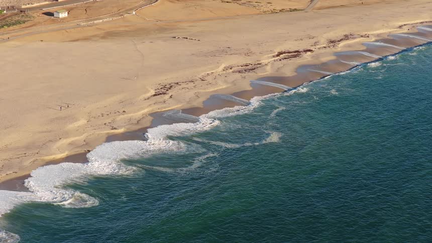 A serene, slow-motion aerial perspective captures the rhythmic motion of turquoise ocean waves washing onto a wide, sandy shore