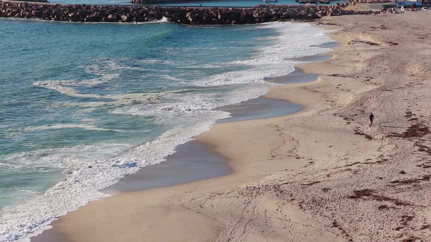 A lone person walks along a wide sandy beach as gentle waves roll ashore in slow motion, with a harbor in the background