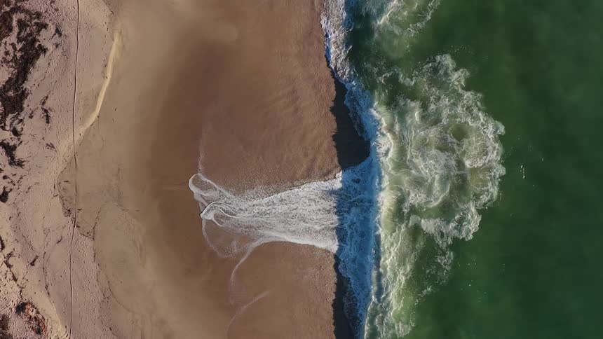 A mesmerizing top-down aerial view shows gentle ocean waves washing onto a sandy beach in slow motion