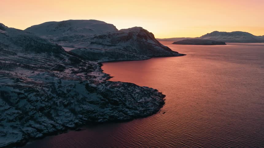 A tranquil aerial view shows a stunning winter sunset casting a warm glow over a snow-covered mountain coastline