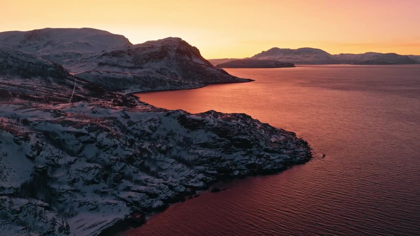 The warm glow of sunset reflects on the calm waters of a fjord surrounded by snow-covered mountains