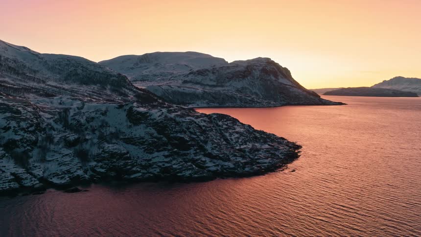 A serene aerial shot captures a tranquil fjord with snow-covered mountains bathed in the soft glow of a colorful winter sunrise