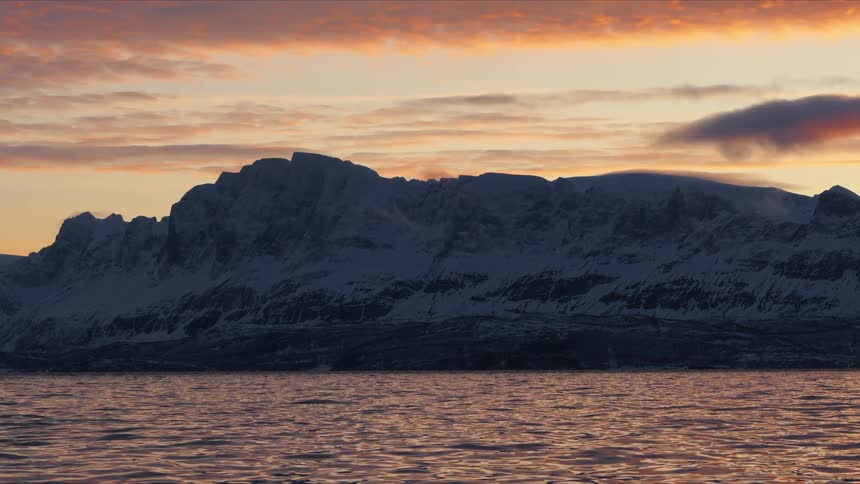 The serene waters of a fjord reflect the warm glow of a winter sunset behind a dramatic snow-covered mountain range