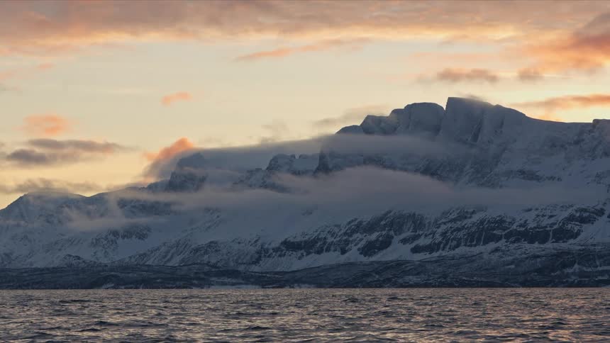 A majestic snow-covered mountain range is shrouded in clouds over a tranquil sea at sunset