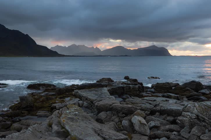 A dramatic sunset breaks through stormy clouds over a rugged mountain coastline and the ocean