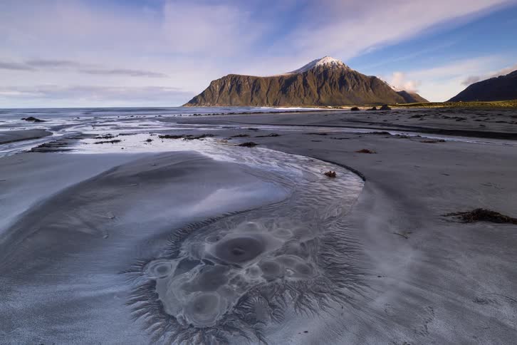 A time-lapse of an otherworldly beach in Lofoten, Norway featuring unique sand patterns, a flowing stream, and a majestic snow-capped mountain