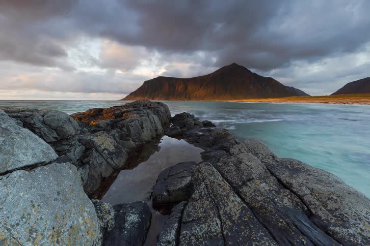 A stunning time-lapse shows golden sunrise light breaking through dramatic clouds to illuminate a majestic mountain across a rocky ocean coastline