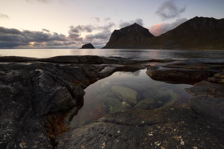 A stunning timelapse captures the changing light of sunset over a rugged, mountainous coastline, with a calm tide pool reflecting the dramatic sky