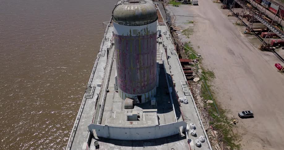 An aerial shot slowly rises over the large, decaying funnels of an abandoned ocean liner docked in a busy industrial shipyard