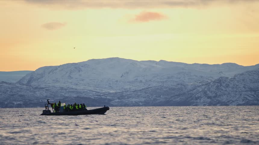 Tourists on a rigid inflatable boat float on the arctic sea with a stunning backdrop of snow-covered mountains at sunset