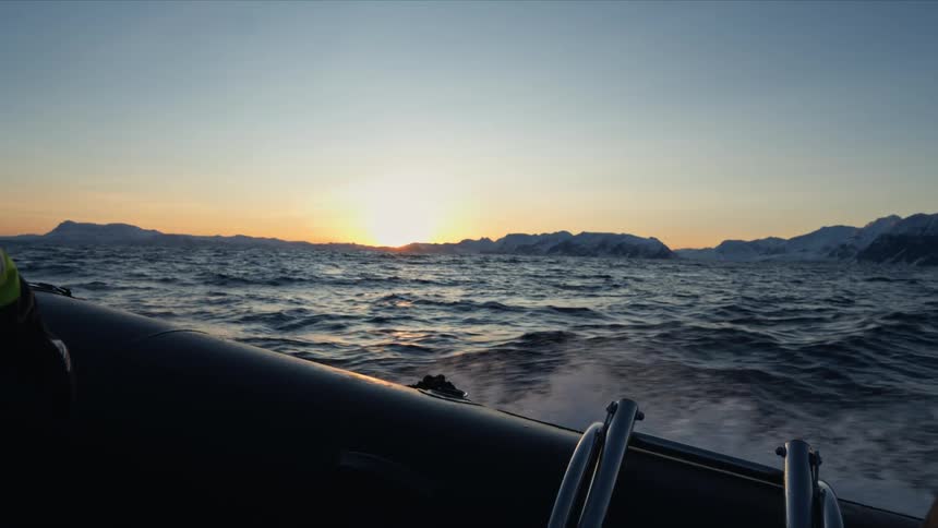A boat speeds across the arctic ocean at sunset, kicking up dramatic plumes of water spray in slow motion against the snowy mountain scenery