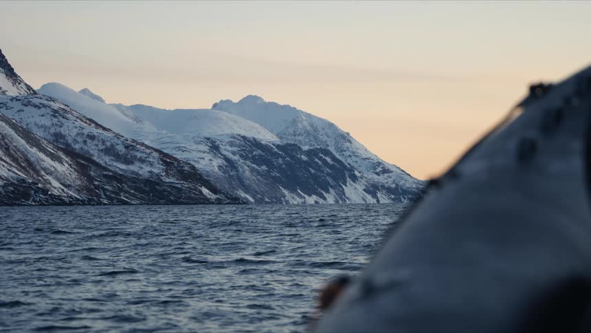 A serene view from an inflatable boat reveals a majestic, snow-covered mountain range across the calm water of a fjord at dawn
