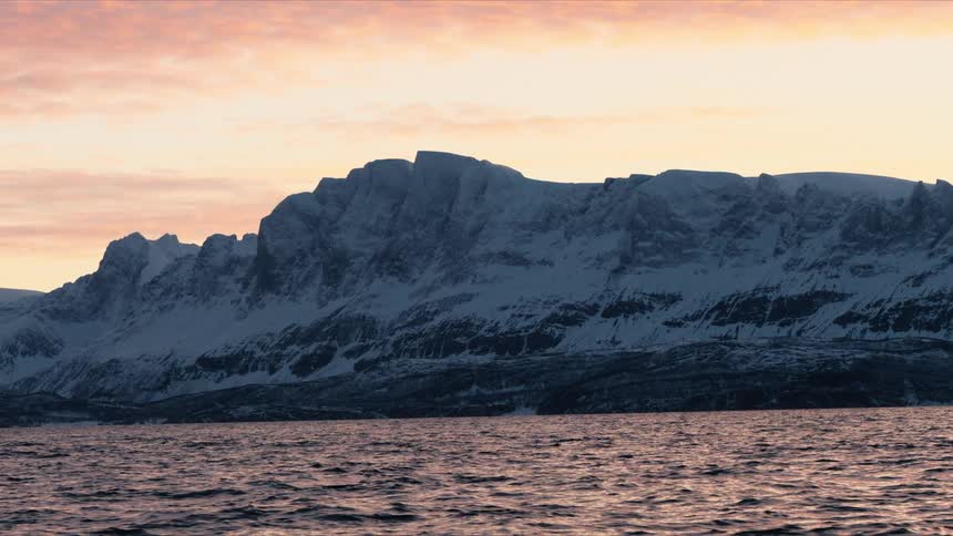 A beautiful winter sunset illuminates a majestic snow-covered mountain range across a tranquil fjord
