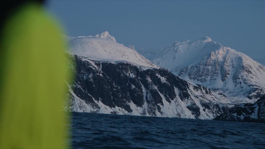 A majestic, snow-covered mountain range rises from the deep blue ocean as seen from a moving boat at dawn