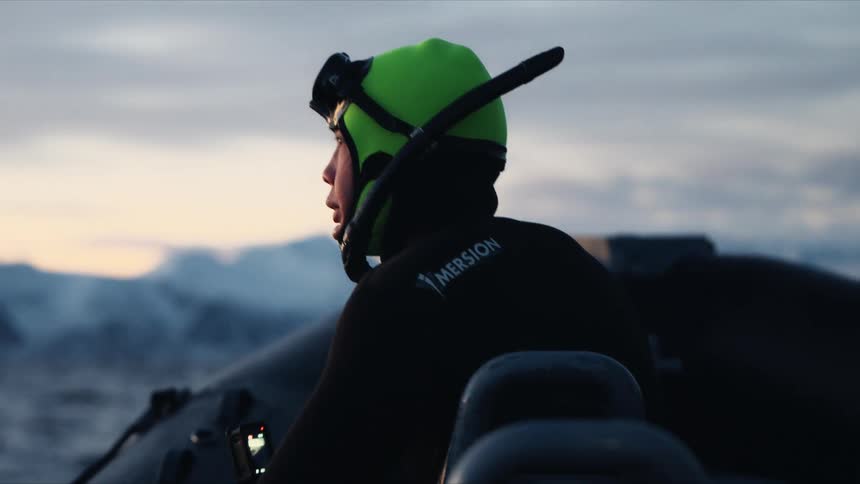 A snorkeler in a wetsuit on a zodiac boat looks out at the snowy arctic mountains before a cold water dive