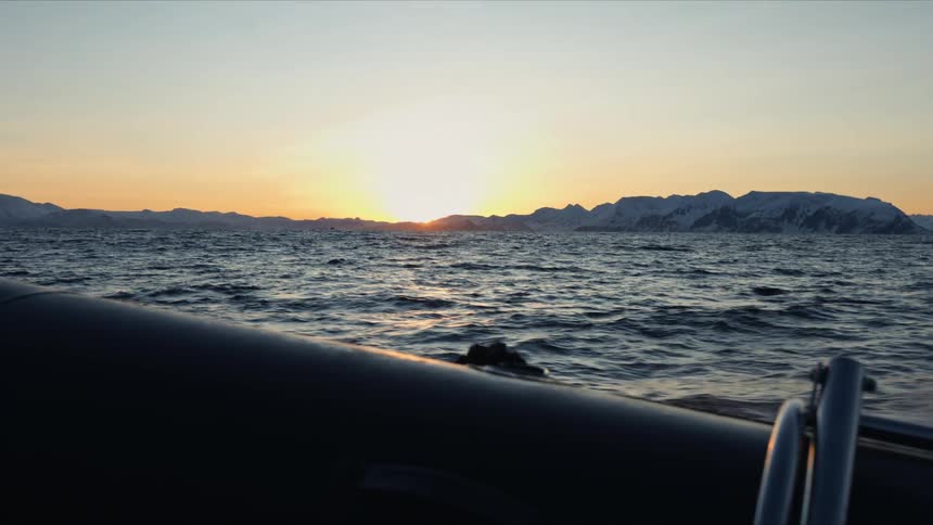 A rigid inflatable boat cruises across the arctic ocean during a golden sunset, with snowy mountains silhouetted against the horizon